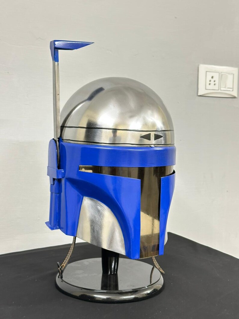 Op de afbeelding: Een blauw en zilverkleurige helm met een kenmerkend ontwerp, met een afgeronde bovenkant en een hoekig vizier. De helm is gemonteerd op een zwarte standaard, die de metallic en glanzende afwerking benadrukt. De helm heeft een zilveren antenne en een blauw accent.