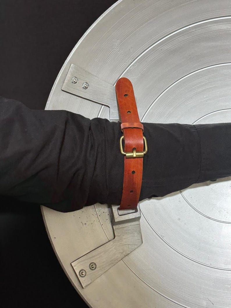 May include: A silver-colored shield with concentric circular ridges. A brown leather strap with a gold-colored buckle is attached to the shield. The shield is held by a person wearing a black long-sleeve shirt.