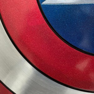 May include: Close-up of a shield with a red, white, and blue color scheme. The shield has a circular design with a silver star in the center. The red sections have a glittery finish.