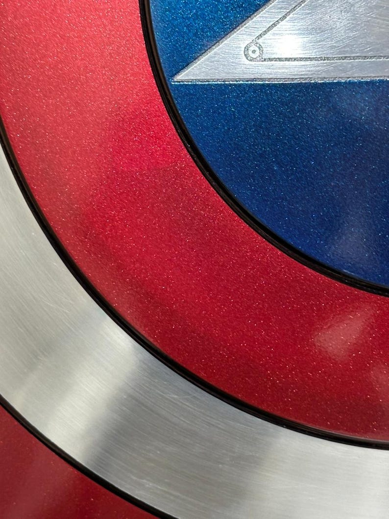May include: Close-up of a shield with a red, white, and blue color scheme. The shield has a circular design with a silver star in the center. The red sections have a glittery finish.