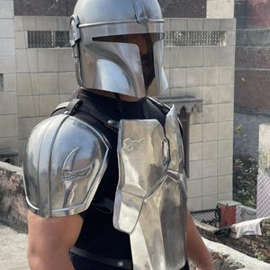 May include: A silver-colored Mandalorian helmet and armor set. The helmet has a T-shaped visor and a rounded top. The armor includes shoulder pads and a chest plate, all with a metallic finish. The set is designed to resemble the iconic look from the Star Wars series.