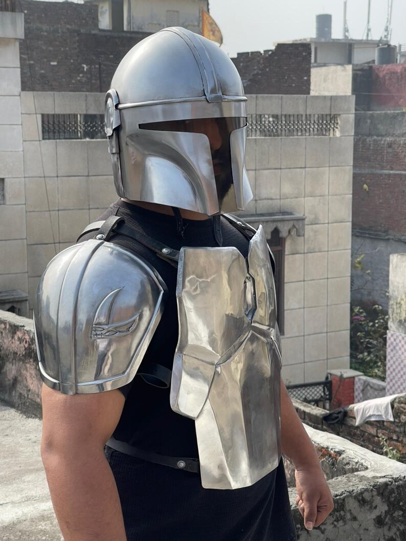 May include: A silver-colored Mandalorian helmet and armor set. The helmet has a T-shaped visor and a rounded top. The armor includes shoulder pads and a chest plate, all with a metallic finish. The set is designed to resemble the iconic look from the Star Wars series.