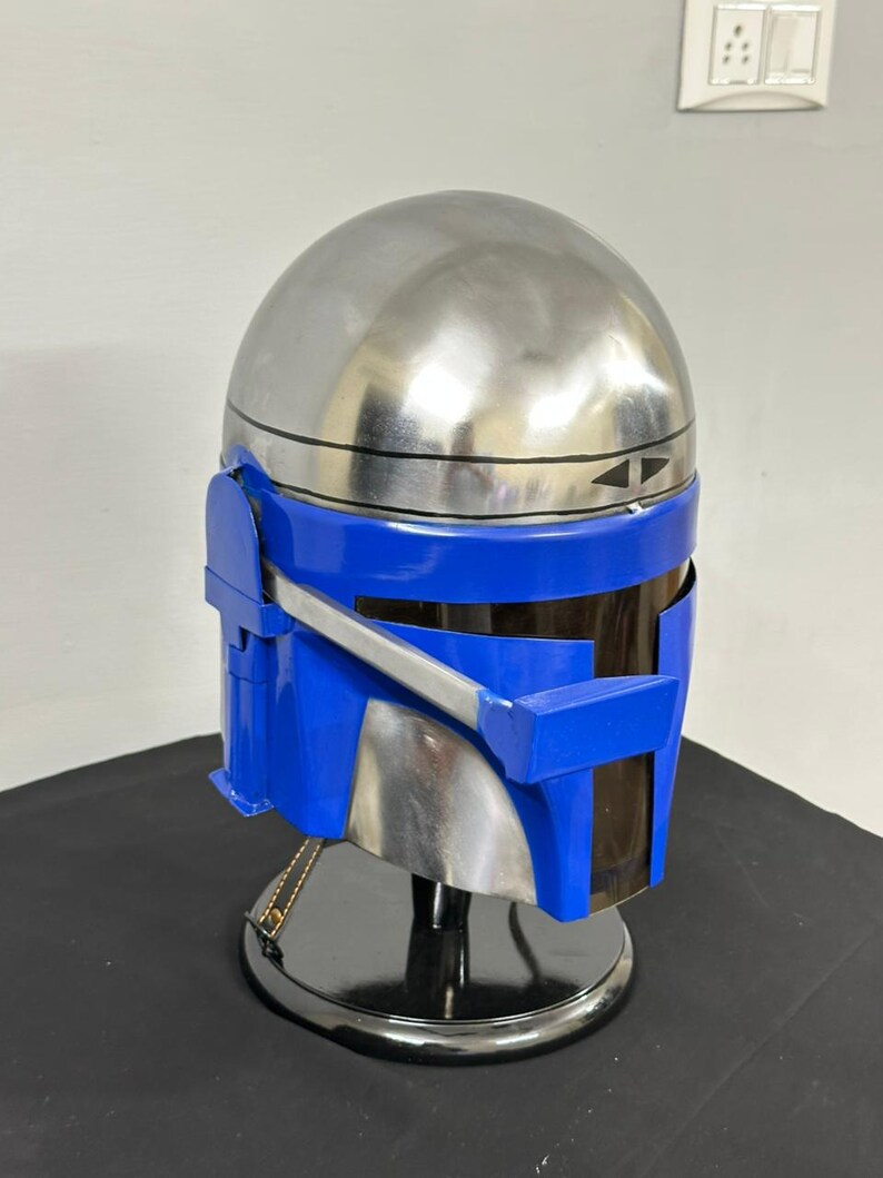 Op de afbeelding: Een zilveren en blauwe helm met een afgeronde bovenkant en hoekige kenmerken. De helm heeft een blauw vizier en accenten, met een reflecterende zilveren afwerking op het hoofdgedeelte. Het wordt tentoongesteld op een zwarte standaard.
