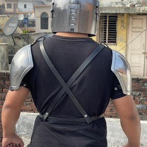 May include: A silver-colored helmet and shoulder armor set with black straps. The helmet has a rounded top and rectangular details. The shoulder armor is curved and metallic. The straps cross over the back and connect at the waist.