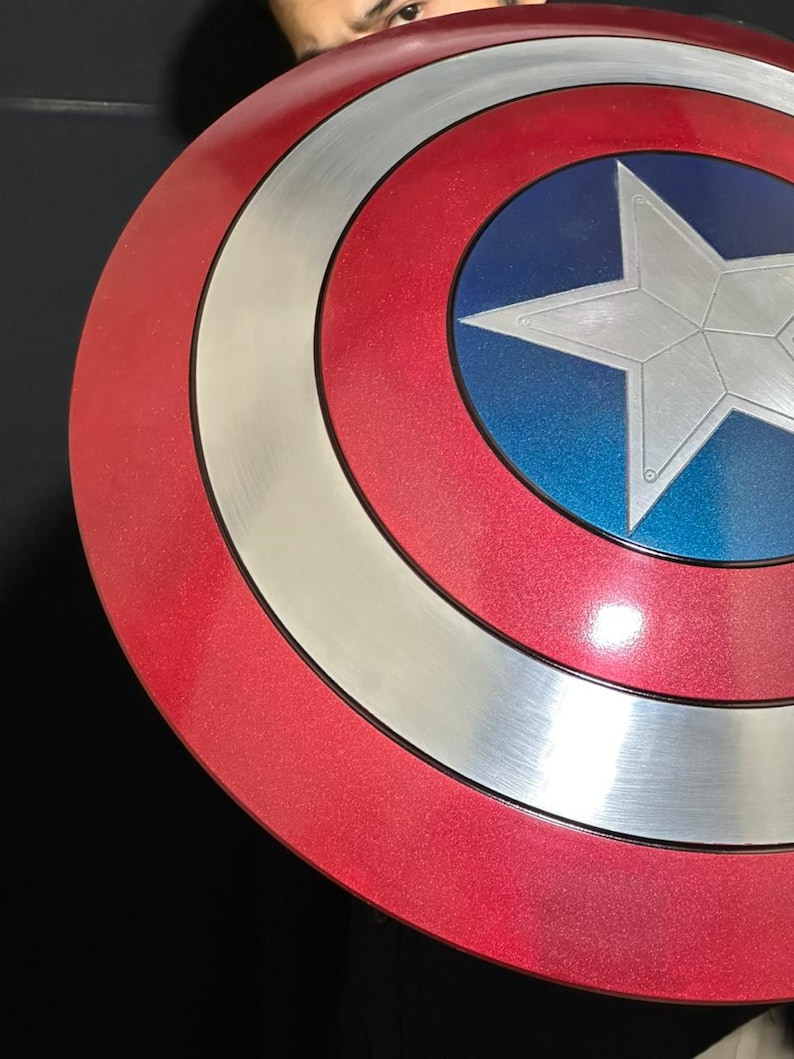 May include: A red, white, and blue shield with a silver star in the center. The shield has a circular design with concentric rings of red, silver, and blue. The shield is a prop or collectible.