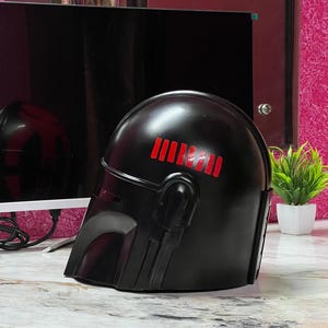 May include: A black helmet with red stripes on top, featuring a visor and side details. The helmet is likely a costume piece or collectible, possibly inspired by a science fiction theme.