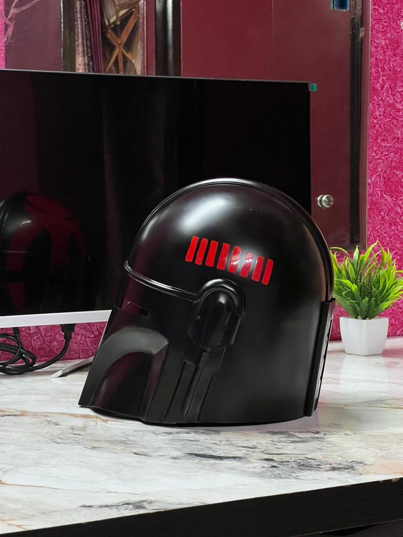 May include: A black helmet with red stripes on top, featuring a visor and side details. The helmet is likely a costume piece or collectible, possibly inspired by a science fiction theme.