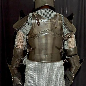 May include: A full suit of medieval armor, viewed from the back. The armor is dark gray and includes a helmet, chest plate, shoulder guards, arm guards, and chainmail. Leather straps and rivets secure the pieces together. The background is black.