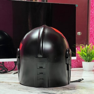 May include: A black helmet with red accents. The helmet has a smooth, glossy finish and features a series of rectangular details on the back. Red markings are visible on the top and side of the helmet. The helmet is sitting on a light-colored surface.