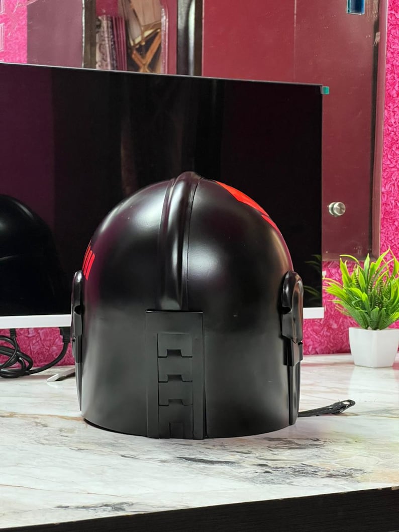 May include: A black helmet with red accents. The helmet has a smooth, glossy finish and features a series of rectangular details on the back. Red markings are visible on the top and side of the helmet. The helmet is sitting on a light-colored surface.