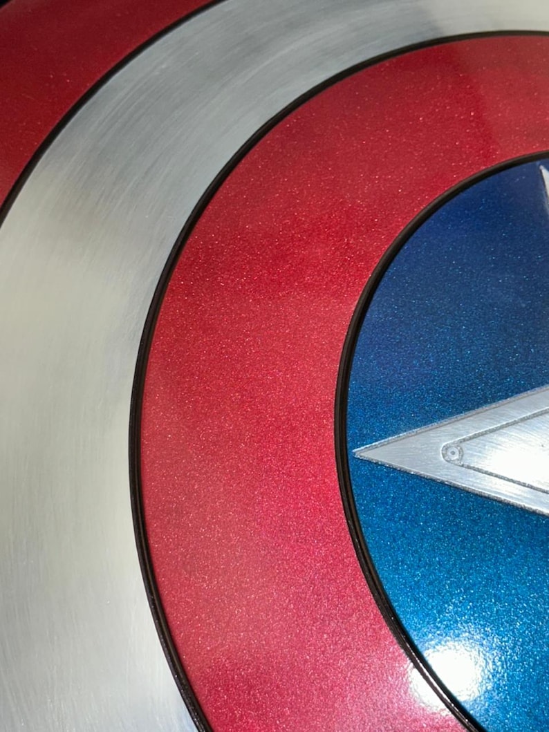 May include: Close-up of a Captain America shield. The shield features a red, white, and blue color scheme with a star emblem. The red sections have a glittery finish, and the shield is outlined in black.