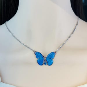 May include: A silver necklace with a blue butterfly pendant. The butterfly pendant has translucent, sparkling wings and a small blue gem. The necklace is displayed on a person's neck.