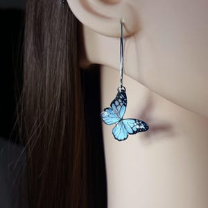 May include: A long, silver-toned earring featuring a dangling, light blue butterfly charm with black vein detailing. The butterfly has a translucent appearance with a subtle iridescent sheen, suspended from a thin, curved silver-toned metal wire.