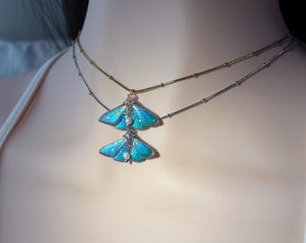 Puede incluir: Un conjunto de collares superpuestos con dos cadenas delicadas, una dorada y otra plateada, cada una adornada con un colgante en forma de mariposa de color azul y verde vibrante. Cada colgante está acentuado con una pequeña perla blanca y cristales.