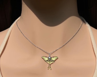 Puede incluir: Un collar de cadena plateada con un colgante en forma de polilla con alas de esmalte amarillo pálido y verde, acentuado por una pequeña piedra preciosa transparente en el centro del cuerpo, suspendido de una delicada cadena de eslabones metálicos.