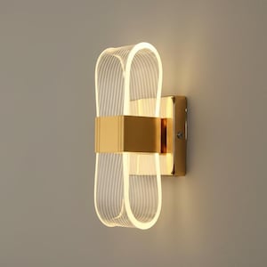 Op de afbeelding: Een moderne wandlamp met een goudkleurige rechthoekige basis en een decoratief, verlicht ontwerp van helder acryl. Het acrylelement heeft een lusvorm met verticale lijnen, waardoor een lintachtig uiterlijk ontstaat. De lamp is op een neutraal gekleurde muur gemonteerd.