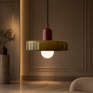 Op de afbeelding: Een moderne hanglamp met een rode bovenkant en een brede, olijfgroene kap. De lamp heeft een bolvormige, witte lamp aan de onderkant en hangt aan een zwart snoer. De armatuur hangt in een kamer met een marmeren vloer en neutraal getinte muren.