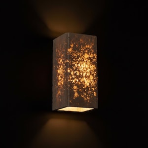 Peut inclure: Applique murale rectangulaire avec une surface tachetée et texturée. L'applique émet une lumière chaude et dorée, créant un contraste avec le fond sombre. Le design est moderne et minimaliste, adapté à l'éclairage intérieur.