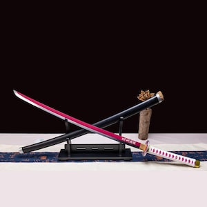 Puede incluir: Una katana con una hoja roja y un mango blanco con un diseño de diamantes. La espada se exhibe sobre un soporte negro con una vaina negra. La hoja tiene un borde plateado y un centro rojo.