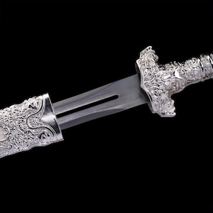 May include: A silver-colored sword featuring a metallic blade with a central groove. The hilt and the top portion of the scabbard are decorated with intricate, raised scrollwork patterns. The metal surfaces exhibit a polished, reflective finish.