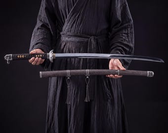 Puede incluir: Una espada katana japonesa con una hoja curva, sostenida horizontalmente. La espada tiene un mango negro con elementos decorativos y una guarda plateada. La vaina es marrón con detalles plateados y borlas. La persona lleva una túnica negra.