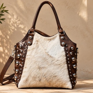 May include: A handbag featuring a central panel of white and tan hair-on hide leather flanked by dark brown leather sides. The brown leather sections are decorated with rows of polished silver-tone metal studs. It includes two top handles and a shoulder strap.