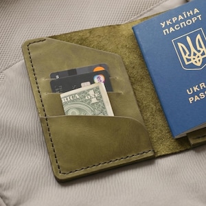 Könnte beinhalten: Olivenfarbener Leder-Reisepasshalter mit blauem ukrainischen Reisepass. Der Halter ist offen und zeigt Kartenfächer und einen US-Dollar-Schein. Der Reisepass hat goldfarbene Schrift.
