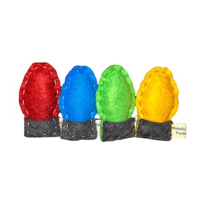 May include: A string of four felt Christmas lights in red, blue, green, and yellow. The lights are attached to a gray felt base. The tag reads "MadeByPortia".