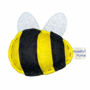 May include: A yellow and black felt bee with white felt wings. The bee has a stitched design and a tag that reads "MadeBy1 Portland".