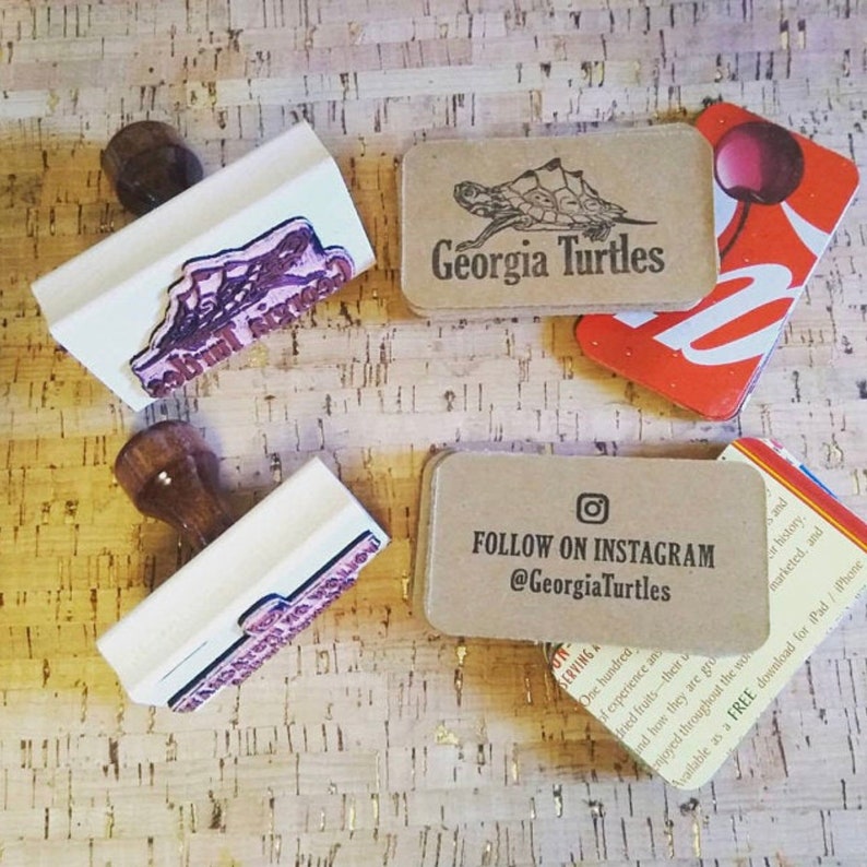 May include: Four brown cardboard business cards with white text. The top card says "Georgia Turtles" and features a black and white illustration of a turtle. The second card says "FOLLOW ON INSTAGRAM @GeorgiaTurtles". The third card is red with white text. The fourth card is partially visible and has white text on a yellow background.