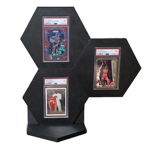 May include: Black hexagonal display case holding three graded basketball trading cards. The cards feature images of basketball players. The display case has a built-in stand for tabletop or shelf display.