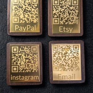 May include: Four wooden QR code plaques with gold-coloured metal plates. Each plate has a unique QR code and the words "Paypal", "Etsy", "Instagram", and "Email" engraved. The plaques are on a dark, textured surface.
