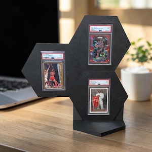 May include: Black hexagonal display stand holding three graded sports cards. The cards feature basketball players in action. The stand is made of a matte black material and is designed to showcase collectible cards.