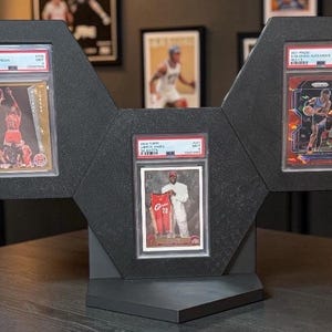 May include: A black, geometric display stand holding three encased sports trading cards. The cards feature basketball players in action. The stand is made of a dark material and is designed to showcase the cards.