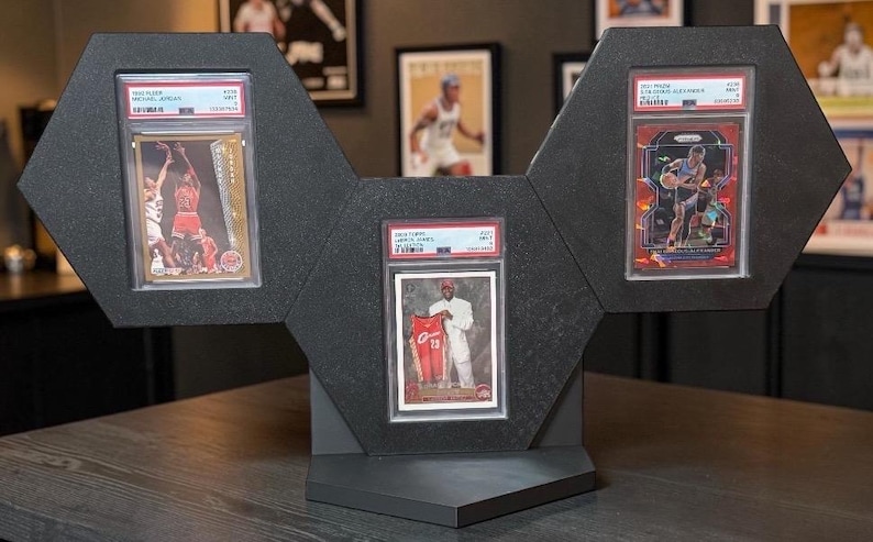 May include: A black, geometric display stand holding three encased sports trading cards. The cards feature basketball players in action. The stand is made of a dark material and is designed to showcase the cards.
