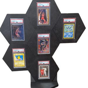 Op de afbeelding: Zwart zeshoekig display voor beoordeelde sport- en ruilkaarten. Het display biedt plaats aan zes kaarten, waaronder een Michael Jordan-kaart, een Pokemon-kaart en andere basketbal kaarten. De behuizing heeft een ingebouwde plank.