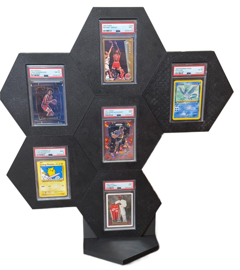 Op de afbeelding: Zwart zeshoekig display voor beoordeelde sport- en ruilkaarten. Het display biedt plaats aan zes kaarten, waaronder een Michael Jordan-kaart, een Pokemon-kaart en andere basketbal kaarten. De behuizing heeft een ingebouwde plank.