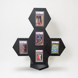Op de afbeelding: Zwarte zeshoekige displaystandaard met vijf beoordeelde ruilkaarten. De standaard is gemaakt van zwart materiaal en heeft een uniek geometrisch ontwerp. De kaarten bevatten afbeeldingen van basketballers en een Pok&eacute;mon.