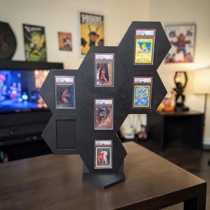 Op de afbeelding: Zwarte zeshoekige displaystandaard voor beoordeelde ruilkaarten. De standaard bevat verschillende kaarten in beschermende hoesjes, met sport- en Pok&eacute;mon-kaarten. De standaard is gemaakt van hout en staat op een tafel.
