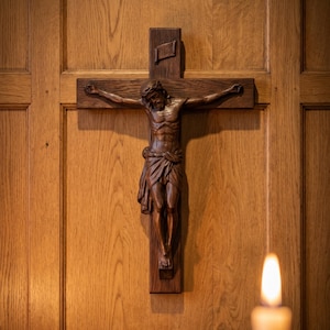 May include: A carved wooden crucifix depicting Jesus Christ on a cross, mounted on a wooden panel. The figure is detailed with a crown of thorns and a loincloth. A lit candle is in the foreground.