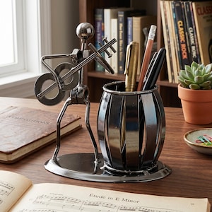 Op de afbeelding: Een metalen bureau-organizer met een muzikant die gitaar speelt. De sculptuur is gemaakt van metalen moeren en bouten. Een pennenhouder is aan de basis bevestigd en bevat pennen en potloden. Een notitieboek en bladmuziek staan op de voorgrond.