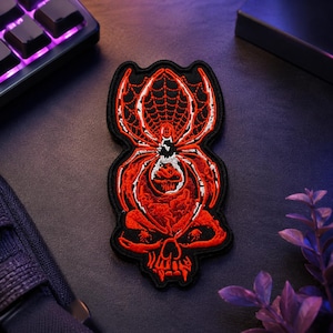 May include: An embroidered patch featuring a red and white spider design centered above a red skull. The spider is depicted with a web pattern on its abdomen, and the entire graphic is outlined in black thread against a black fabric background.
