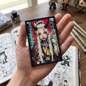 Op de afbeelding: Een rechthoekige patch met een zwarte rand, met een afbeelding van een vrouw met hoorns, tatoeages en lang wit haar. Het kunstwerk heeft een rode en turquoise achtergrond met Japanse tekst.