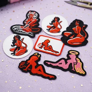 May include: A selection of embroidered patches with red devil designs. The patches show a devilish figure with horns and a tail in different poses, set against black and white backgrounds. The patches are in various shapes, including round and rectangular.