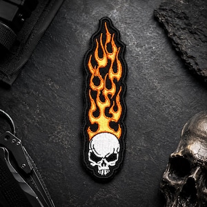 May include: A black embroidered patch featuring a white skull at the base with orange and yellow flames rising upwards. The patch has a black border and is designed for application on clothing or accessories.