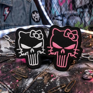 May include: Two black embroidered patches featuring a skull design with a bow. One patch has white detailing, the other has pink. The patches are on a leather surface with safety pins and a chain.