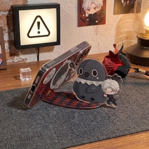 May include: A clear phone case with a red and black design is propped up by a clear stand featuring a cartoon bird and character. The stand sits on a gray surface. A small light with an exclamation point is in the background.