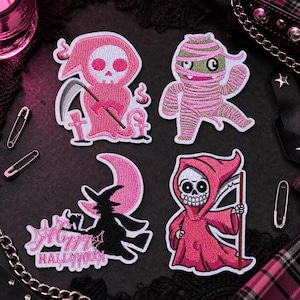 以下が含まれることがあります： ハロウィーンをテーマにした4つの刺繍パッチ。ピンクの死神、ピンクのミイラ、三日月を持つ魔女、鎌を持った死神が描かれています。1つのパッチには「Happy Halloween」の文字があります。
