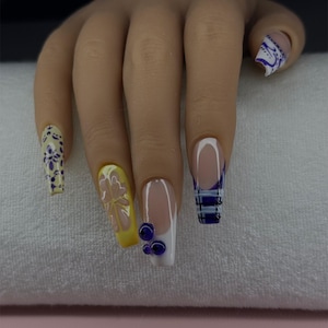 May include: A hand with long, square-shaped acrylic nails. The nails feature various designs, including a French tip with blue stripes, yellow with a flower, and a floral pattern. Some nails have 3D blue embellishments.