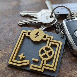 May include: A gray and gold keychain featuring an electrical circuit design with a lightning bolt symbol. The keychain is attached to a keyring with keys and a car key fob.